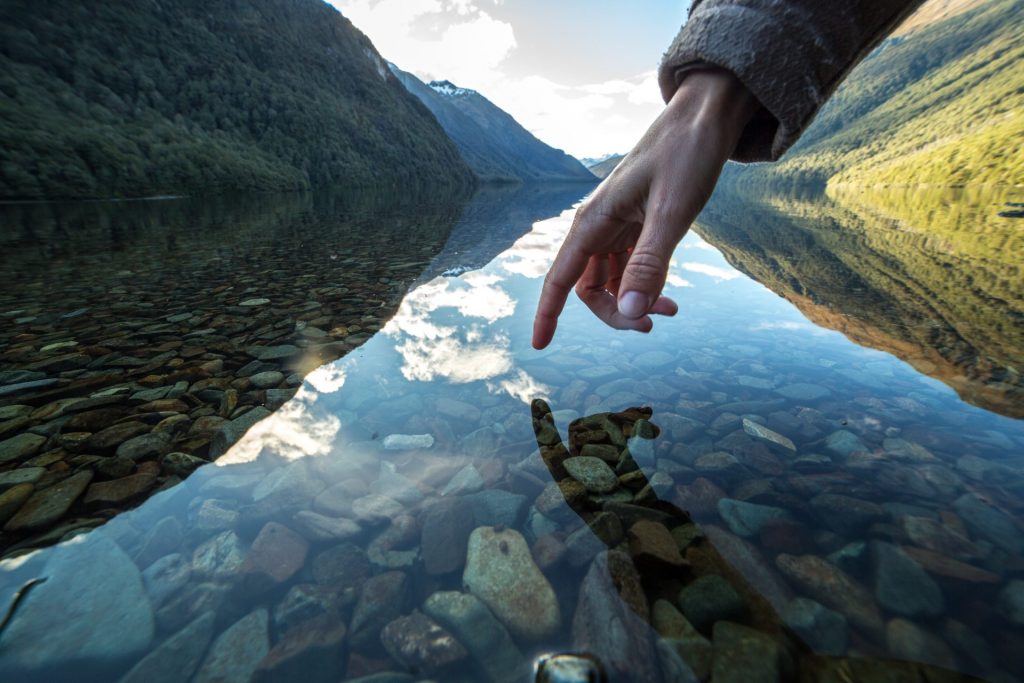 Finger touches surface of mountain lake, the landscape is reflecting on the water.