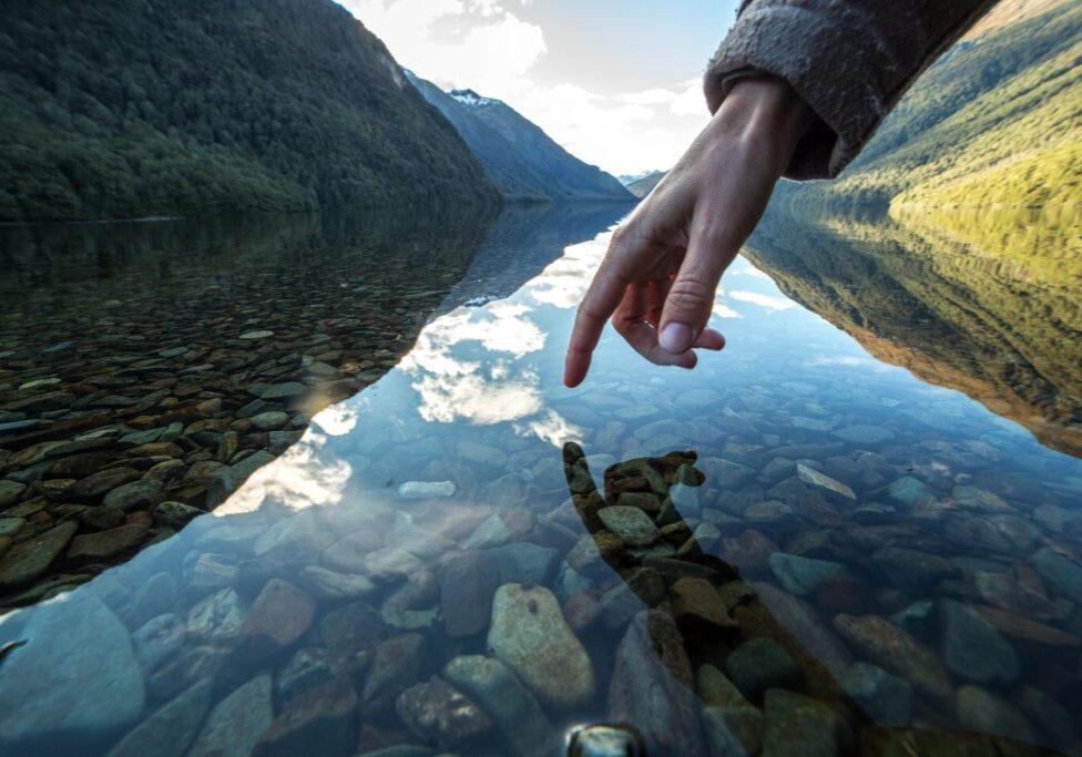 Finger touches surface of mountain lake, the landscape is reflecting on the water.